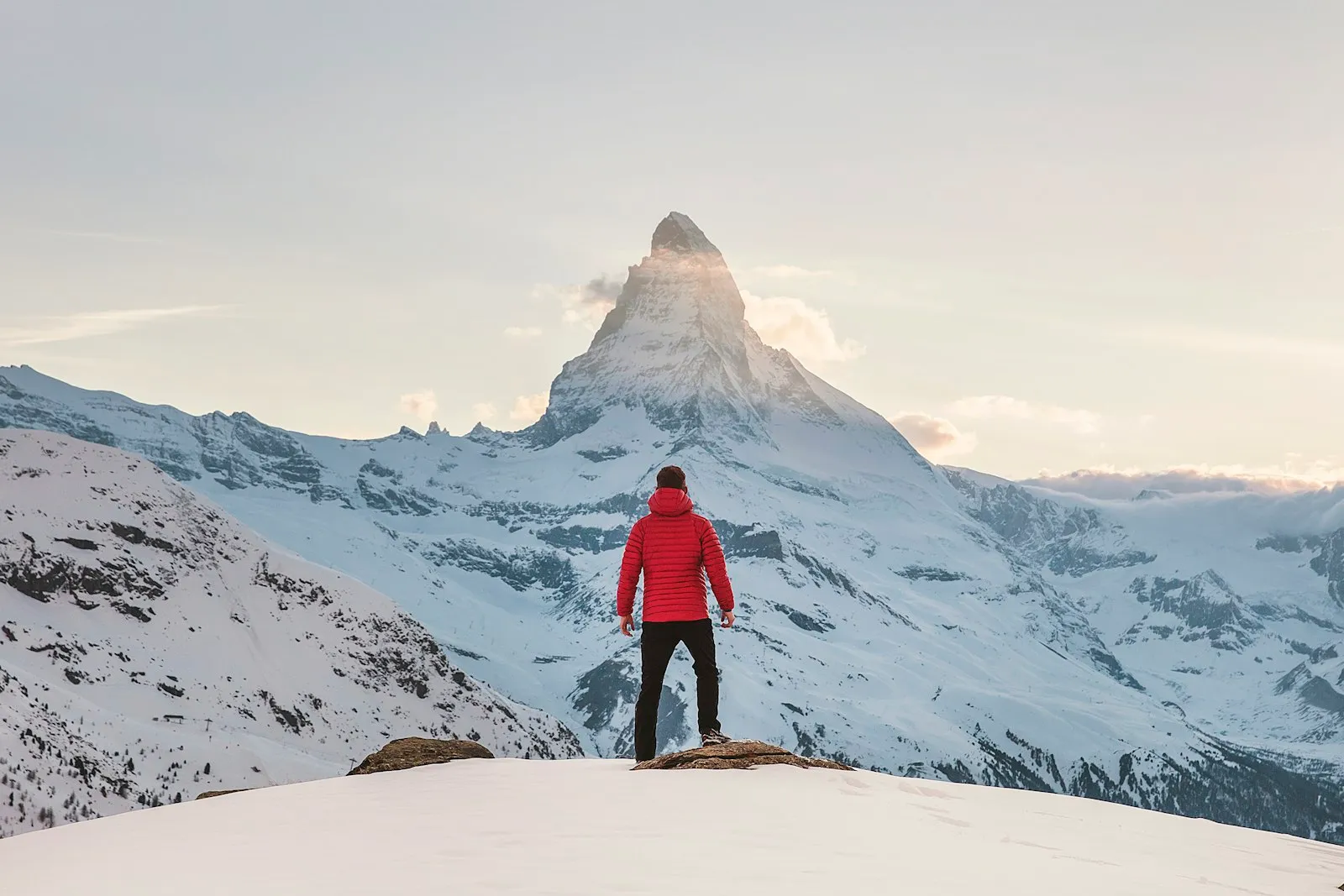 A person in a red jacket standing on a snowy ridge looking at a peak